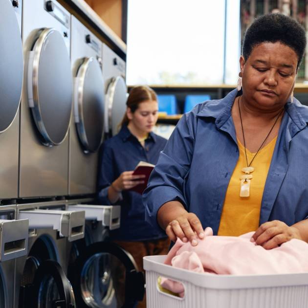 Medium shot of senior African American woman sorting through laundry in basket on folding table, while preparing to load clothes into washing machine at coin operated laundromat