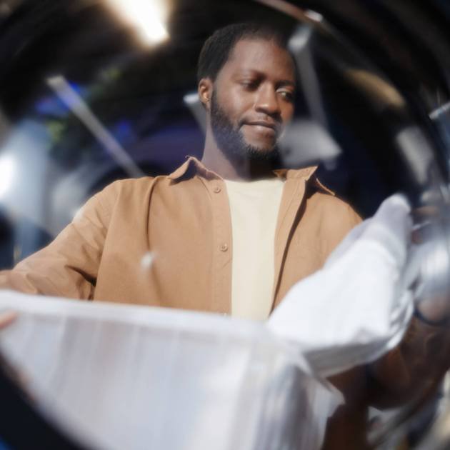 Young African American man with plastic basket containing clothes standing behind open door of washing machine in laundry cafe